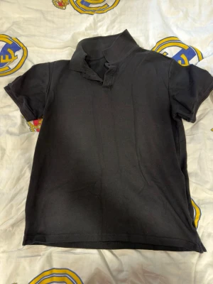 Svart kortärmad polotopp - En stilren svart polotopp med korta ärmar och klassisk krage. Toppen har knappar vid halsen och är gjord i ett mjukt material som känns bekvämt mot huden. Perfekt för en enkel och clean look.