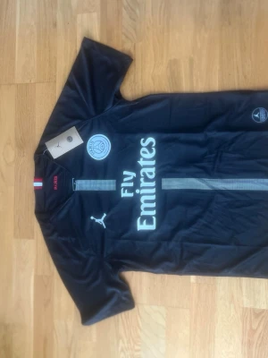 PSG Neymar Jr matchtröja svart - Snygg svart PSG matchtröja med Neymar Jr och nummer 10 på ryggen. Tröjan har Fly Emirates-tryck på bröstet, Paris Saint-Germain-logga och Puma-logga. Korta ärmar och fransk flaggdetalj i nacken. Perfekt för fotbollsfans!