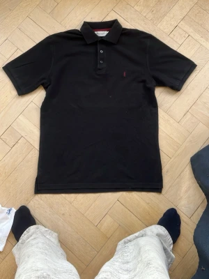 Svart pikétröja från Yves Saint Laurent - Klassisk svart pikétröja från Yves Saint Laurent med krage, tre knappar och diskret röd logga broderad på bröstet. Tröjan har korta ärmar och är tillverkad i ett mjukt bomullsmaterial som känns skönt mot huden.