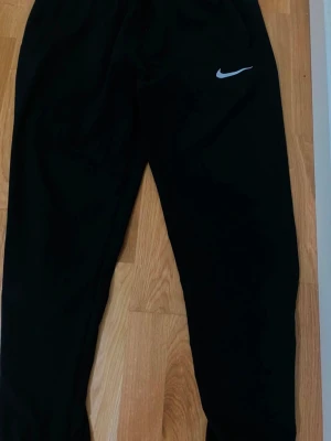 Svart Nike Miller  - Säljer ett svart träningsset från Nike med byxor och jacka. Båda plaggen har den klassiska vita Nike-loggan. Jackan har huva och dragkedja framtill samt fickor med dragkedja. Byxorna har resår i midjan och är i ett lätt material, perfekt för träning.