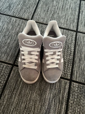 Grå Adidas sneakers med mocka - Snygga grå sneakers från Adidas med klassiska vita ränder på sidorna och stor logga på plösen. Skorna är i mocka med vita skosnören och innerfoder. Perfekta för dig som gillar retrostil och streetwear.