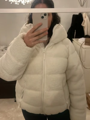 Vit teddy pufferjacka med huva - Supermysig vit pufferjacka i teddy-material med dragkedja framtill och två fickor med dragkedja. Jackan har en hög krage och huva. Puffig och oversized passform. Den håller en verkligen jättevarm och den säljs ej längre❣️köpt för 1100kr