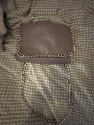 Grå axelremsväska med kedjedetaljer - Snygg grå handväska från Tiamo i konstläder med silverfärgade kedjedetaljer runt kanterna och som axelrem. En liten defekt. Se bild 3