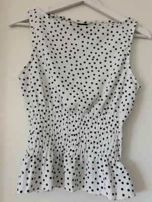 Vit prickig topp från Gina Tricot - Säljer en vit ärmlös topp från Gina Tricot med svarta prickar. Toppen har smock i midjan och volang nedtill, vilket ger en snygg och feminin siluett. Materialet är lätt och stretchigt, perfekt för varma dagar.