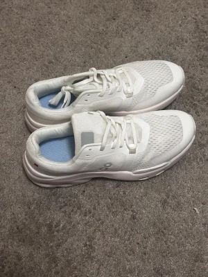 Vita sneakers från On Running - Stilrena vita sneakers från On Running med ventilerande mesh-ovandel och klassisk snörning. Skorna har en låg profil och diskreta detaljer i grått samt en bekväm, vadderad insida med ljusblått foder. Perfekta för en sportig och clean look.