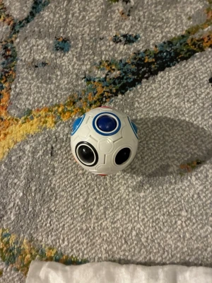 Fidgetboll med färgade knappar - En vit fidgetboll med flera färgade knappar i blått, svart, rött, lila, grönt och fler färger. Bollen har små hexagonmönster och varje knapp är omgiven av en ring i matchande färg. Perfekt för att sysselsätta händerna och minska stress. Den röda ringen är borta. Annars bra skick!!