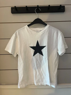 Vit t-shirt med svart stjärna - Vit t-shirt med en stor svart stjärna tryckt på bröstet. Klassisk rund halsringning och korta ärmar. Perfekt för en avslappnad och trendig stil. Materialet känns mjukt och bekvämt, troligtvis bomull.