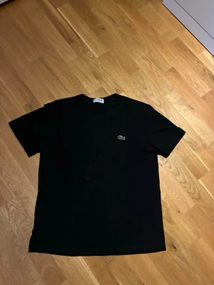 Lacostet-shirt - Klassisk svart t-shirt från Lacoste med den ikoniska krokodilloggan broderad på bröstet. T-shirten har rund hals och korta ärmar, perfekt för en clean och stilren look. Tillverkad i mjuk bomull för maximal komfort.