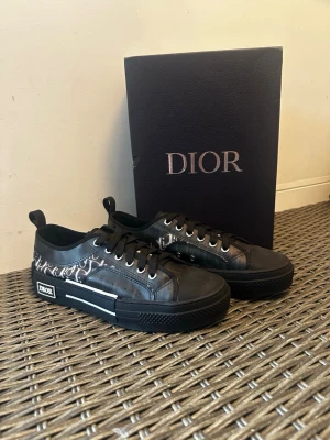 Svarta Dior b23 sneakers - Snygga svarta Dior sneakers med vit logotypdetalj på sidan och svart sula. Skorna har lågt skaft, svarta skosnören och en stilren design med monogrammönster längs sidan. Perfekta för dig som vill ha en exklusiv och trendig look.