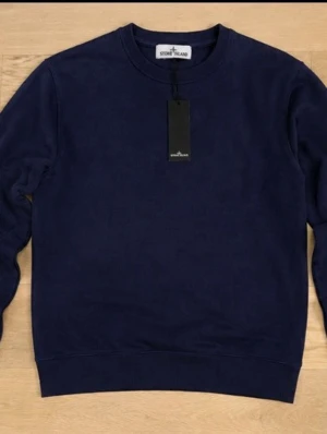 Mörkblå sweatshirt från Stone Island - Mörkblå sweatshirt från Stone Island med klassisk rund halsringning och ribbade muddar. Tröjan har den ikoniska Stone Island-loggan på vänster ärm. Perfekt för en clean och stilren look. Passar även S.