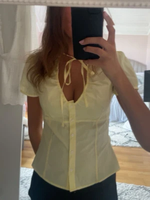 Zara Gul blus med knyt och puffärm - Söt ljusgul blus med korta puffärmar och knytband vid halsen. Blusen har knappar framtill och figurnära passform som framhäver midjan. Perfekt för en trendig och feminin look.