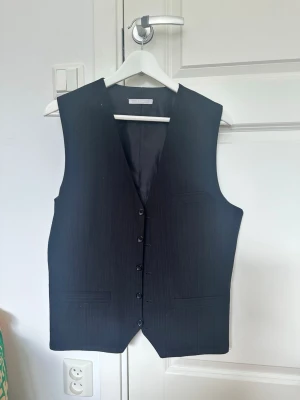 Svart randig väst från These Glory Days - Klassisk svart väst med tunna vita ränder från These Glory Days. Västen har knäppning framtill, två fickor och en stilren, rak passform. Perfekt för att lyfta din outfit med en dressad touch.