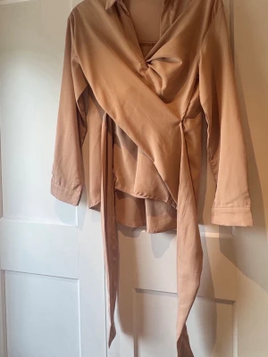 Beige/rosa  omlottblus med långa ärmar - Snygg beige omlottblus med långa ärmar och knytband i midjan. Blusen har en klassisk krage och ett mjukt, följsamt tyg som ger en elegant look. Perfekt att styla med jeans eller kjol för en trendig outfit.