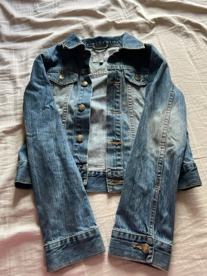 Klassisk blå jeansjacka - Snygg blå jeansjacka med slitna detaljer och silverfärgade knappar. Jackan har två bröstfickor med lock och knapp samt långa ärmar med knapp vid ärmslut. Perfekt att slänga över en hoodie eller t-shirt för en avslappnad look.