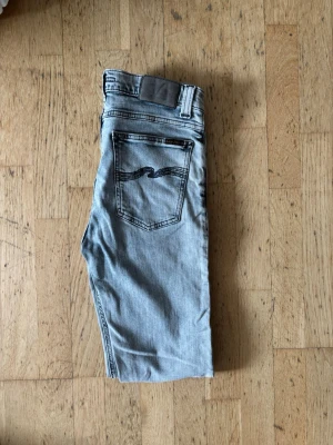 Nudie jeans - Säljer ett par ljusblå jeans med smal passform och klassisk femficksdesign. Jeansen har en snygg bakficka med vågig detaljsöm och bälteshällor. Perfekta för en avslappnad och trendig look. Materialet är mjukt denimtyg.