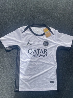 PSG Tröja (Helt Ny) - Säljer en vit PSG tröja från Jordan med svarta detaljer och Dri-FIT-material. Tröjan har Paris Saint-Germain-logga, Jordan-logga och Qatar Airways-tryck på bröstet. Perfekt för fotboll eller träning.