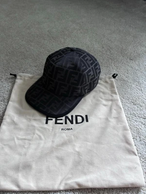Svart keps med Fendi-logga - Säljer en svart keps från Fendi med det klassiska FF-mönstret över hela kepsen. Kepsen har böjd skärm och justerbar rem bak. Perfekt accessoar för dig som vill ha en lyxig och trendig look.