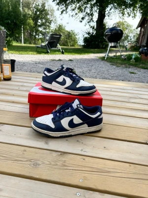 Nike Dunk Low blå och vita sneakers - Säljer ett par Nike Dunk Low sneakers i blått och vitt. Skorna har klassisk låg siluett, vit tåbox och sidor med mörkblå overlays och matchande blå snören. Ikoniska Nike Swoosh-loggan på sidan och vit mellansula. Perfekta för dig som gillar streetwear och sportig stil. Använda i 2 månader har lite slitningar