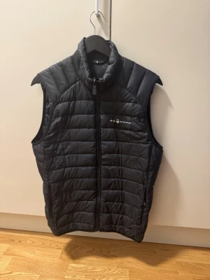 Svart dunväst från Sail Racing - Snygg svart dunväst från Sail Racing med quiltad design och hög krage. Västen har dragkedja framtill och logga på bröstet. Perfekt för lager-på-lager och håller dig varm under kyliga dagar.