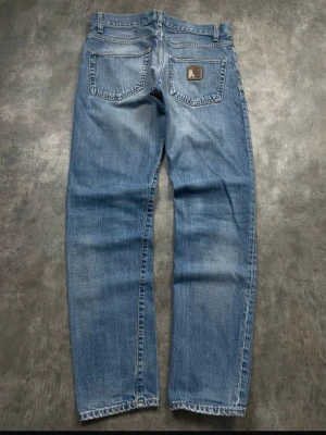 Carhartt jeans - Säljer ett par klassiska blå jeans från Carhartt med rak passform och fem fickor. Jeansen har en snygg tvättad look och Carhartt-lapp på bakfickan. Tillverkade i slitstark denim, perfekta för en avslappnad streetwear-stil.