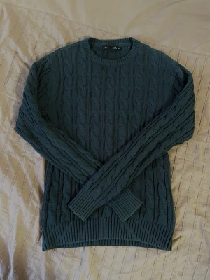 Mörkgrön kabelstickad tröja - Säljer en mörkgrön kabelstickad tröja med rund halsringning och ribbade muddar vid ärmslut och nederkant, vilket ger en klassisk look. Perfekt att ha över en skjorta! Tröjan har dock krympt lite pga. tvätt, så ha i åtanke att den kan sitta lite mindre än M. Den är gjord av 100% bomull.  Axelbredd: 45 cm.   Längd: 71 cm.