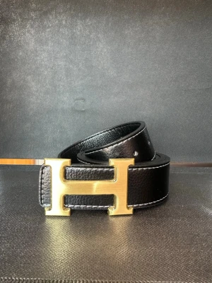 Svart skärp med guldfärgat H-spänne - Snyggt svart bälte i skinn med ett stort guldfärgat H-format spänne. Bältet har en stilren design med vita sömmar längs kanterna och passar perfekt till jeans eller kostymbyxor. Ett statement-plagg för dig som gillar klassiska accessoarer.