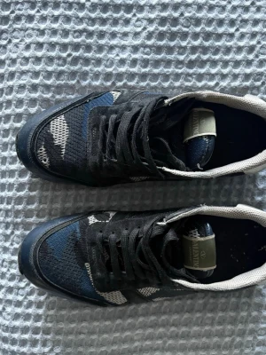 Valentino sneakers med blå kamouflage - Säljer ett par snygga Valentino sneakers med svart och blå kamouflagemönster. Skorna har snörning, rund tå och detaljer i textil och skinn.