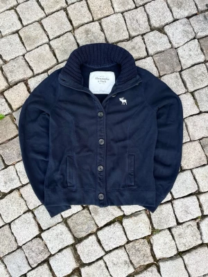 Mörkblå Cardigan från Abercrombie & Fitch - Mörkblå kofta med ribbad, hög krage och knappstängning framtill. Klassisk logga med älg broderad på bröstet. Två fickor fram och mjukt material som känns skönt mot huden. Perfekt för lager på lager och en stilren look. L i barnstorlek, ungefär S i vanlig storlek.
