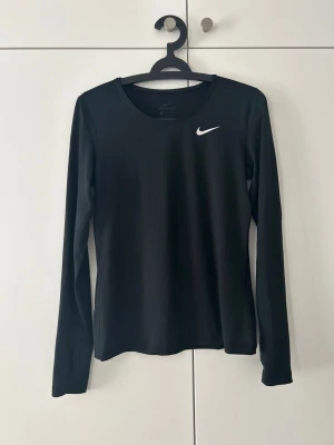 Svart långärmad träningströja Nike - Svart långärmad träningströja från Nike med vit logga på bröstet. Tillverkad i ett stretchigt funktionsmaterial som andas, perfekt för löpning eller gym. Tumhål i ärmsluten för extra komfort. Snygg och enkel design som passar till all träning.