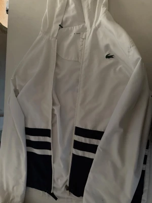 Lacoste jacka - Snygg vit vindjacka från Lacoste med marinblå ränder och klassisk krokodil-logga på bröstet. Jackan har huva, dragkedja framtill och elastiska muddar vid ärmslut och midja. Perfekt för en sportig och stilren look. Som ni kan se på dom 3 sista bilderna har jackan små fläckar som inte går att få bort. Däremot är inte fläckarna inte jätte synliga.