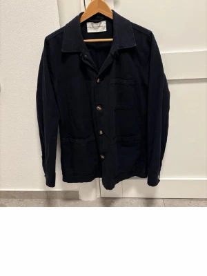 Mörkblå overshirt med fickor - Stilren mörkblå overshirt med klassisk krage och fyra framfickor. Overshirten har knäppning framtill med stora knappar och är tillverkad i ett kraftigare bomullstyg. Perfekt lager-på-lager-plagg för en avslappnad look.