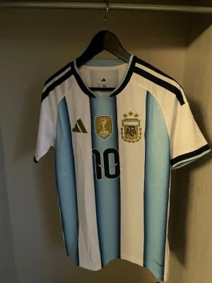 Argentina Messi fotbollströja - Säljer en officiell Argentina fotbollströja från Adidas med Messi och nummer 10 på ryggen. Tröjan har klassiska blå och vita ränder, svarta detaljer och FIFA/AFA-emblem på bröstet. Perfekt för dig som älskar fotboll och vill stötta landslaget.