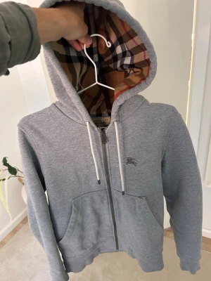 Grå zip hoodie från Burberry - Snygg grå zip hoodie från Burberry med klassiskt rutigt foder i huvan och broderad logga på bröstet. Hoodien har vita dragsnören, två fickor fram och är tillverkad i mjuk bomullsblandning. Perfekt för en chill och stilren look.