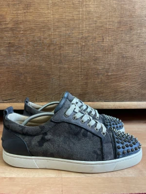 Christian Louboutin  - Unika svarta sneakers med gråvit sula och vita skosnören. Ovandelen är i mockaliknande material och tån är täckt av coola metallnitar som ger en edgy look. Perfekta för dig som vill sticka ut och addera lite attityd till din outfit.