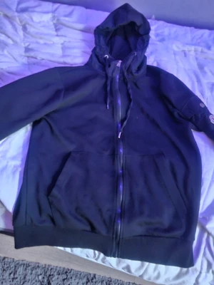 Svart hoodie med dragkedja från Moncler - Svart hoodie från Moncler med dragkedja framtill, huva med dragsko och klassisk logotyp på ärmen. Hoodien har känguruficka och ribbade muddar. Materialet är mjukt och bekvämt, perfekt för en avslappnad stil.