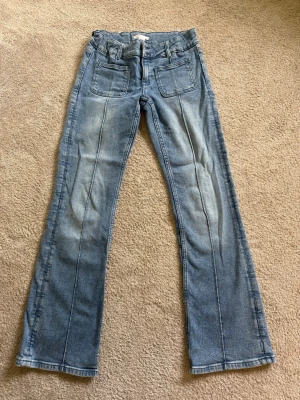 Blå bootcut jeans med fickor - Säljer ett par ljusblå bootcut jeans med framfickor och klassiska bakfickor. Jeansen har markerade sömmar längs benen och är tillverkade i denim.