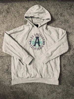 Grå hoodie med tryck Los Angeles - Säljer en grå hoodie med huva och magficka. På bröstet står det 'Los Angeles California Champs' i blått och grönt tryck tillsammans med siffrorna 1978. Hoodien har ribbade muddar och är i mjukt sweatshirtmaterial.