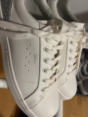 Vita sneakers med glitterdetalj - Fräscha vita sneakers från ONLY med klassisk låg profil och snörning. Skorna har en snygg silverglittrig detalj på hälen och perforerade hål på sidan för extra stil. Aldrig använda🩷