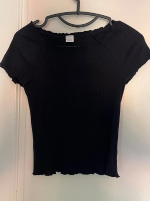 Svart ribbad topp med vågig kant - En svart ribbad topp med korta ärmar och vågiga kanter vid hals, ärmslut och nederkant. Toppen har en tight passform och är tillverkad i ett stretchigt material som sitter snyggt på kroppen. Perfekt att styla med jeans eller kjol.