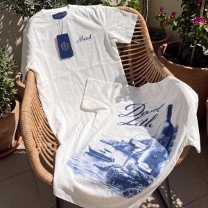 Vit t-shirt med blått tryck - Vit t-shirt med blått tryck på ryggen som föreställer vinflaska, glas och havsmotiv. Framsidan har en liten blå text vid bröstet. Klassisk passform och rund halsringning. Perfekt för en avslappnad och stilren look.
