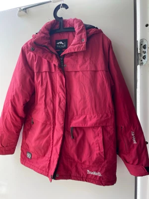 Röd parkas från TrueNorth - Röd parkasjacka från TrueNorth med huva och dragkedja framtill. Jackan har stora fickor, justerbar huva och logga på ärmen samt framtill. Perfekt för kalla dagar och outdoor-äventyr. Materialet är slitstarkt och vattenavvisande.