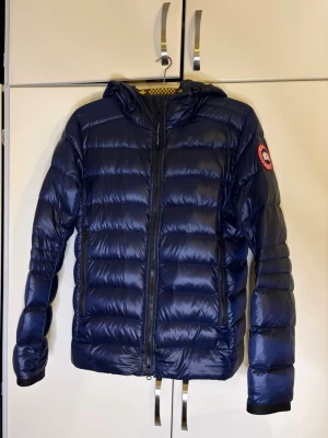 Mörkblå dunjacka med huva - Snygg mörkblå dunjacka från Canada Goose med huva och dragkedja framtill. Jackan har två sidofickor med dragkedja och den klassiska logotypen på vänster ärm. Perfekt för kalla vinterdagar och riktigt lätt att bära.