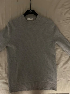 Ljusgrå stickad tröja från Selected - En stilren ljusgrå stickad tröja från Selected med rund halsringning och ribbade muddar vid ärmslut och nederkant. Tröjan har en klassisk rak passform och är tillverkad i ett mjukt, tunt stickat material som passar perfekt till lager på lager.