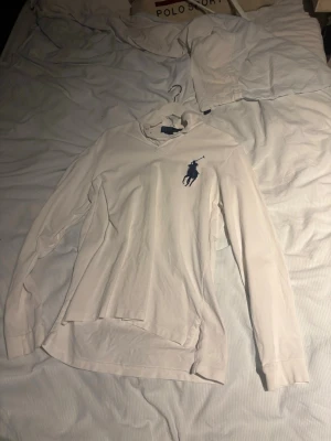 Vit långärmad skjorta Polo Ralph Lauren - Klassisk vit långärmad skjorta från Polo Ralph Lauren med stor mörkblå logga på bröstet. Skjortan har krage och är tillverkad i mjuk bomull, perfekt för en clean och stilren look.