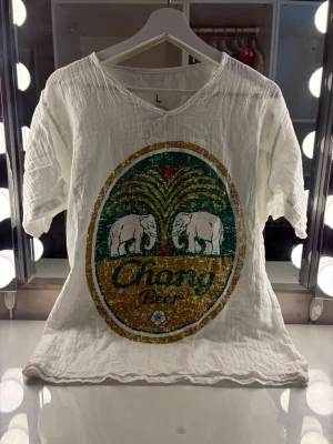 Vit Chang Beer t-shirt med elefanter - Säljer en ikonisk T-shirt med  Chang Beer-tryck framtill i  retrostil. T-shirten har v-ringning och är tillverkad i ett tunt, luftigt bomullsmaterial. Perfekt för en avslappnad sommarlook eller att styla med baggy jeans. T-shirten  har har ett snyggt, lite "slitet" tryck för den rätta vintage-känslan.Perfekt för dig som gillar unika och avslappnade plagg.