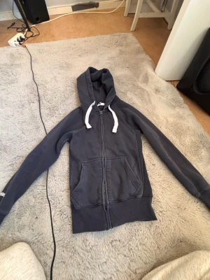 Mörkgrå hoodie med dragkedja - Säljer en mörkgrå hoodie med dragkedja och vita snören i huvan. Tröjan har två stora fickor framtill och ribbade muddar vid ärmslut och nederkant. Perfekt för en avslappnad och chill stil.