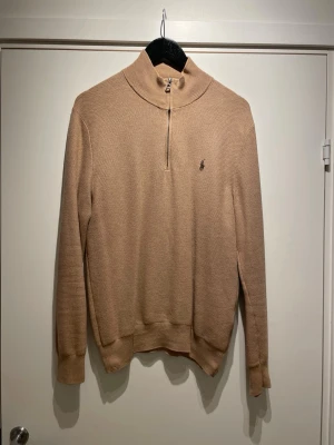 Beige halvzip tröja från Polo Ralph Lauren - Snygg beige stickad tröja från Polo Ralph Lauren med halvzip och hög krage. Tröjan har lång ärm, ribbade muddar och den klassiska broderade loggan på bröstet. Perfekt för lager på lager och en clean look.