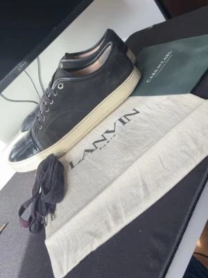Svarta Lanvin sneakers med lackdetalj - Snygga svarta sneakers från Lanvin med klassisk låg profil. Skorna har ovandel i mocka och tåparti i svart lack, samt vita sulor. Kommer med extra skosnören och dustbag. Perfekta för dig som gillar stilrena och exklusiva sneakers.