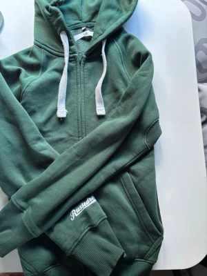 Grön hoodie från Russedress - Mörkgrön hoodie med dragkedja från Russedress. Tröjan har huva med vita snören, ribbade muddar vid ärmslut och nederkant samt fickor på sidorna. Tillverkad i mjukt bomullsmaterial och har Russedress-logga tryckt på ärmen.