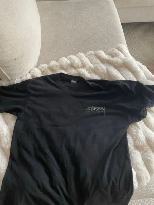 Svart Stüssy t-shirt med logga - Säljer en svart t-shirt från Stüssy med deras ikoniska logga tryckt på bröstet och i större format på ryggen. Klassisk passform och rund halsringning. Perfekt för dig som gillar streetwear och enkel stil. Aldrig använd 